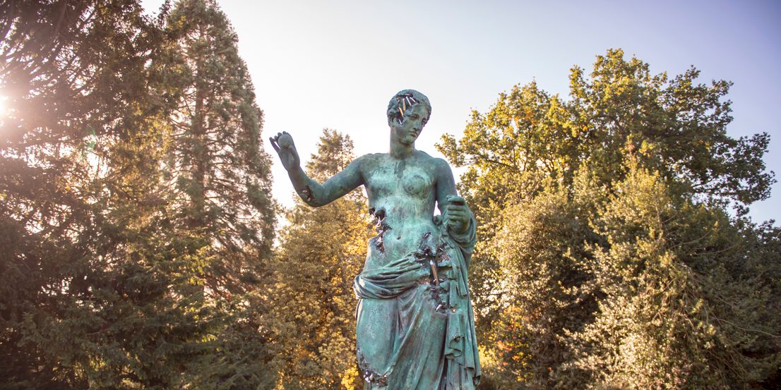 A large bronze sculpture of a woman wearing a draped robe with her arms outstretched.