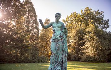 A large bronze sculpture of a woman wearing a draped robe with her arms outstretched.