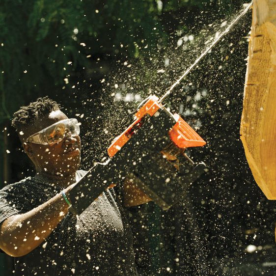 A black woman wearing safety goggles, carving a piece of wood using a chainsaw