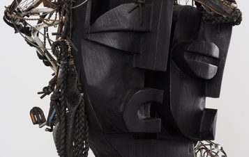 A wooden sculpture of two headed humanoid figure, with hair & accessories made from bicycle parts