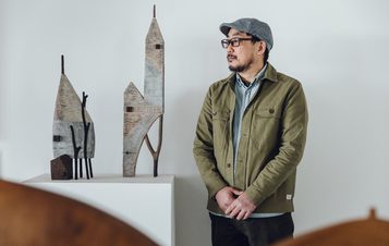 The artist Yukihiro Akama, a Japanese man wearing a green jacket and grey cap, standing next to two carved wooden houses.