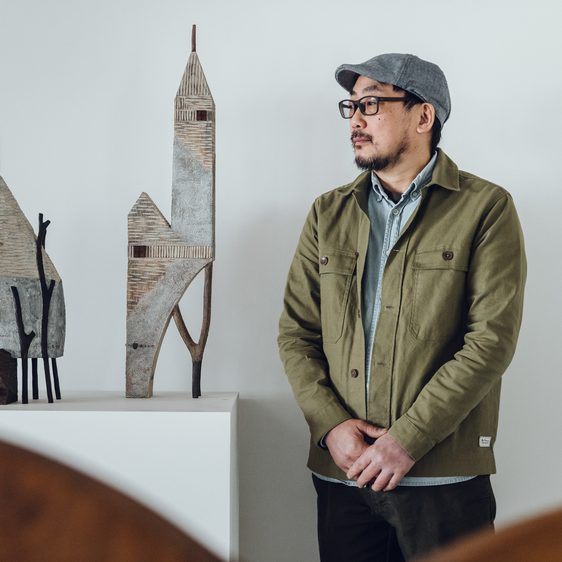 The artist Yukihiro Akama, a Japanese man wearing a green jacket and grey cap, standing next to two carved wooden houses.