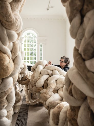 A woman wearing black works on a large installation made from twisting textile forms, in a brightly lit gallery space.