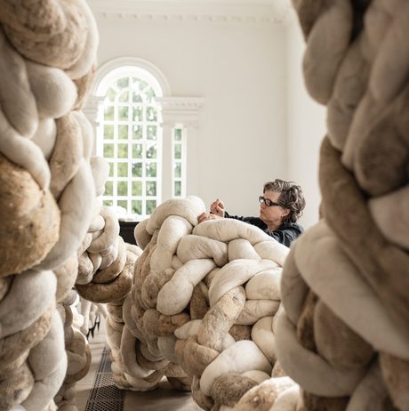 A woman wearing black works on a large installation made from twisting textile forms, in a brightly lit gallery space.