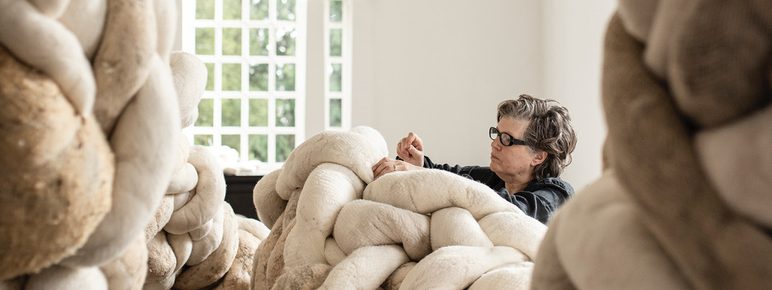 A woman wearing black works on a large installation made from twisting textile forms, in a brightly lit gallery space.