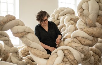 A woman wearing black works on a large installation made from twisting textile forms, in a brightly lit gallery space.