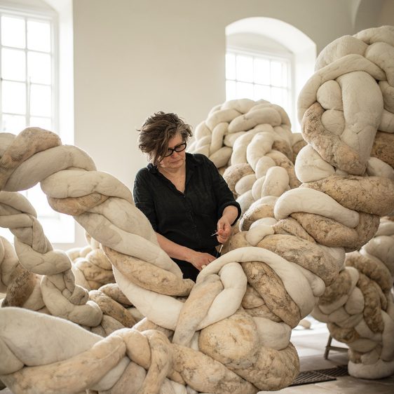 A woman wearing black works on a large installation made from twisting textile forms, in a brightly lit gallery space.