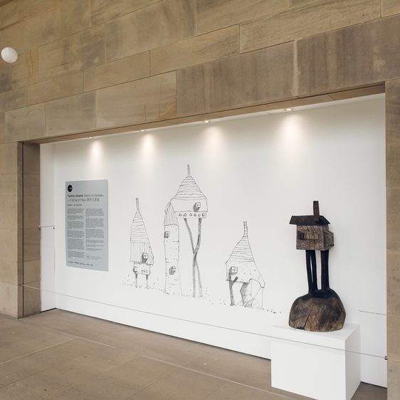 A carved wooden house displayed next to a series of large scale house drawings.