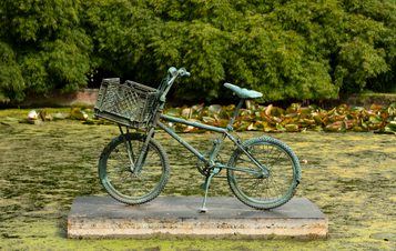 A bronze bicycle mounted on a plinth in the centre of a pond, with bushes and trees in the background.