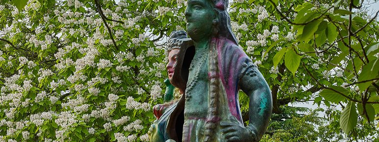 A rainbow coloured statue of an Indian woman surrounded by trees in blossom.