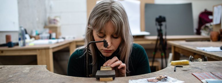 A woman with long blonde hair working on a piece of metal at a workspace.