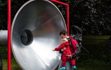 A man and a baby looking into a giant silver tube with a red frame