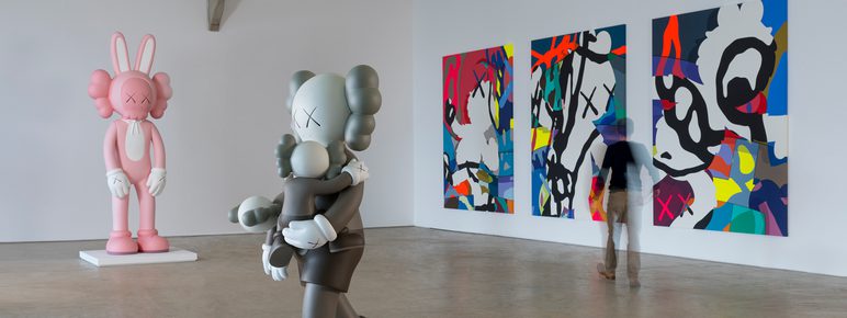 Two large colourful cartoon-style sculptures and wall based artworks in a gallery space. A person can be seen moving between the artworks.
