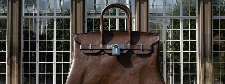 A giant brown Birkin handbag in front of a building with glass windows.