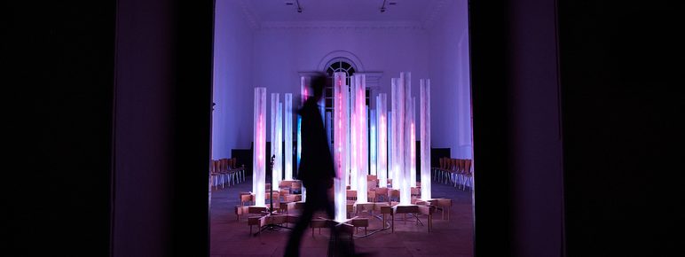 A silhouetted person walking in front of an installation of pink and blue glowing columns.