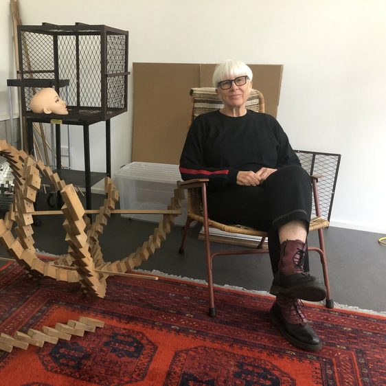 The artist Deborah Rundle sitting in a chair next to a wooden sculpture