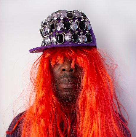 A black man wearing a long red wig and jewel covered baseball cap