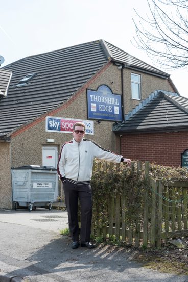 A man wearing a white jacket and sunglasses, standing outside a pub.