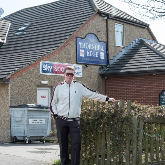 A man wearing a white jacket and sunglasses, standing outside a pub.