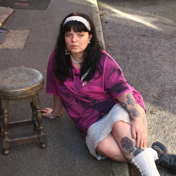 A woman wearing a pink shirt and white skirt, sitting on the ground next to a wooden stool.