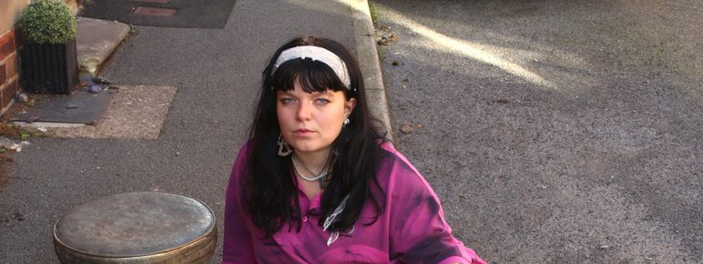A woman wearing a pink shirt and white skirt, sitting on the ground next to a wooden stool.