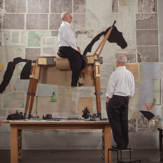 A man looks up at a sculpture of a horse made from found objects, with an image of himself sitting on top of it. Behind them is a wall covered in maps, drawings and texts.