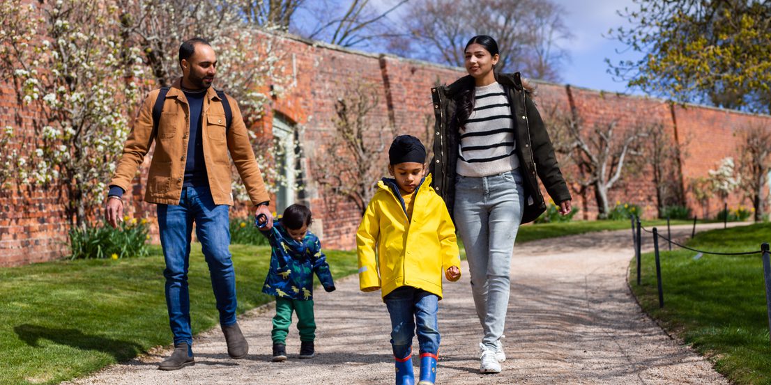 A family of 2 adults and 2 young children, walking along a path. A wall lined with spring blooms is behind them.