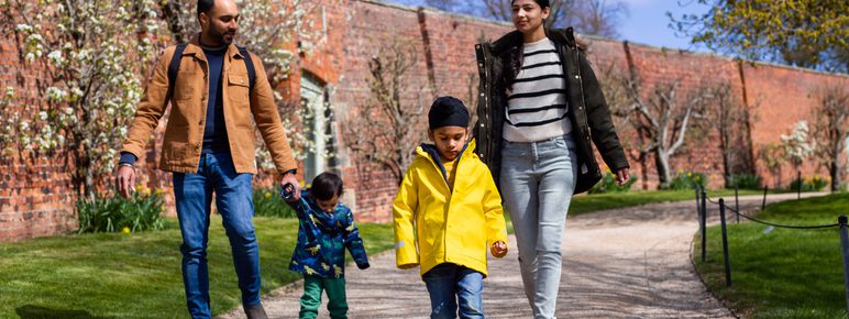 A family of 2 adults and 2 young children, walking along a path. A wall lined with spring blooms is behind them.