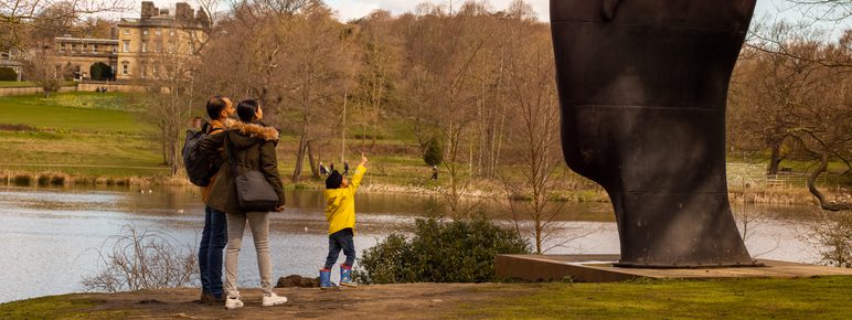 Two adults and a child looking at a giant head sculpture. A manor house and lake is behind them