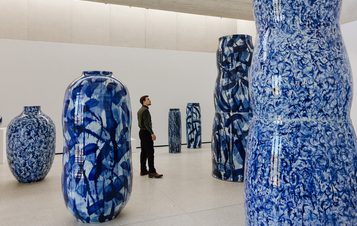 A visitor stands in an art gallery, gazing at a collection of large, intricately designed blue and white vases. The vases vary in shape and size, showcasing abstract patterns against a minimalist white backdrop. Natural light illuminates the space.