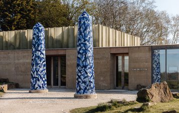 The image shows a modern building named "The Weston," featuring two tall, patterned blue columns at the entrance. The structure has a sleek, metallic roof and large glass windows, surrounded by a natural landscape with trees and rocks.