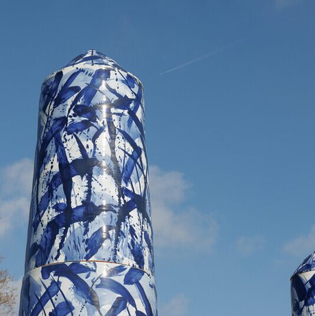 A close-up of two tall, cylindrical sculptures covered in blue and white abstract patterns. They stand against a clear blue sky with a few clouds, and a faint contrail from an aircraft is visible in the upper right corner.