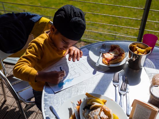 a child drawing at a table in ysp cafe while family are eating lunch outside