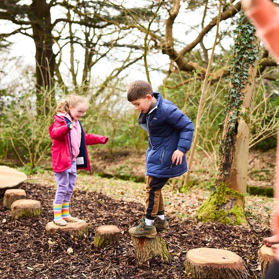 Two children in raincoats playing on wooden stepping stones.