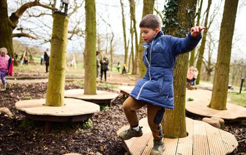 A child jumping from a wooden platform in a woodland play area.