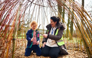 An adult and a child sitting inside a willow den outdoors.