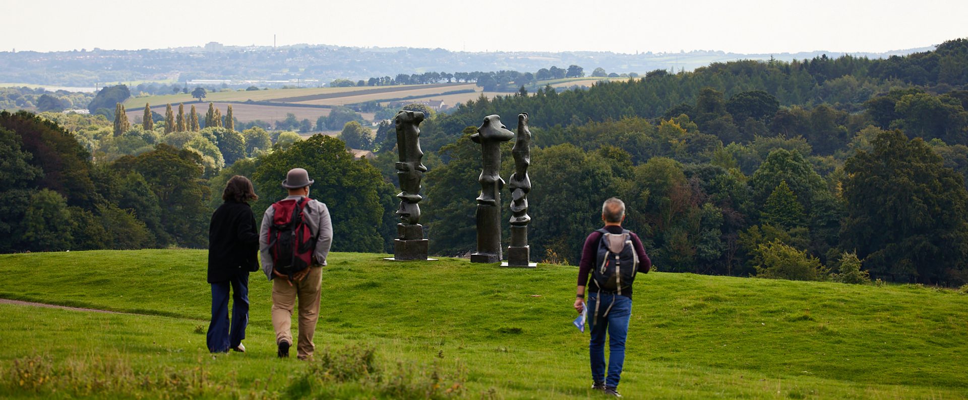 A group of people walking towards three tall thin sculptures on a hillside.