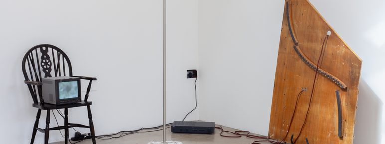 An installation featuring a chair, analogue tv screen, wooden and plaster elements in a white wall gallery space.