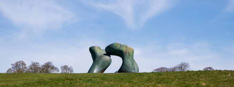 A large abstract bronze sculpture displayed outdoors on a hillside against a blue sky.