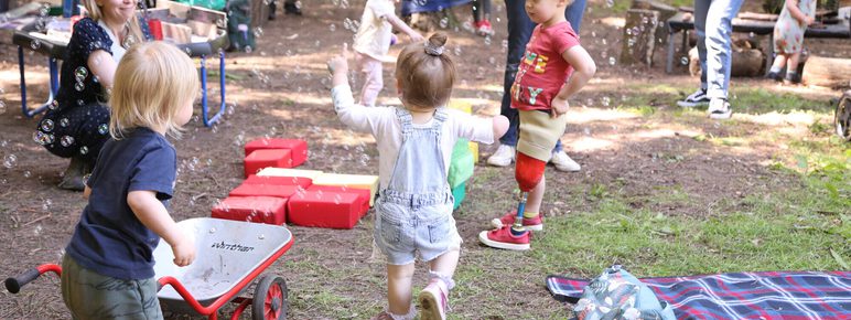 A lively outdoor scene with children playing in a forested area. One child in overalls runs towards the camera, while others play with bubbles. Adults are seated nearby, and toys like a wheelbarrow and picnic blanket are scattered on the grass