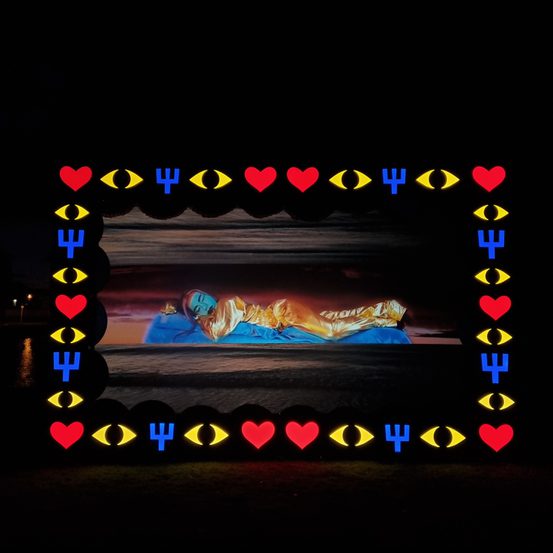 A still from a digital film showing a reclining figure. A custom lightbox frame with motifs of hearts, eyes and cacti surrounds the image.