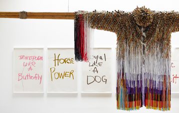 A colourful fringed jacket hangs from a wooden pole. Behind it, framed artworks, each with a different phrase, are displayed on a white wall.