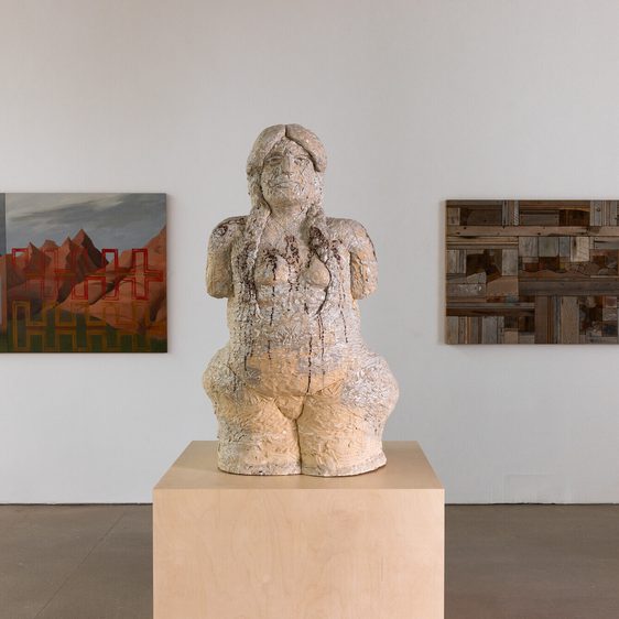 A cream coloured textured sculpture of a human figure on a wooden plinth. Wall based works in shades of brown can be seen on either side.