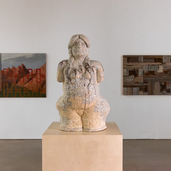 A cream coloured textured sculpture of a human figure on a wooden plinth. Wall based works in shades of brown can be seen on either side.