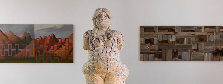 A cream coloured textured sculpture of a human figure on a wooden plinth. Wall based works in shades of brown can be seen on either side.