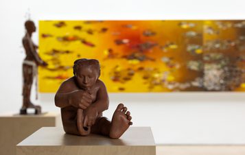 A small clay sculpture of a person building themselves out of raw clay. An abstract painting in shades of orange and a figure sculpture can be seen in the background.