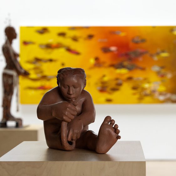 A small clay sculpture of a person building themselves out of raw clay. An abstract painting in shades of orange and a figure sculpture can be seen in the background.