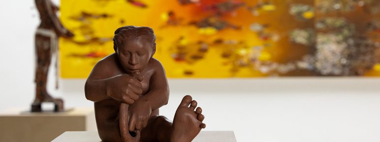 A small clay sculpture of a person building themselves out of raw clay. An abstract painting in shades of orange and a figure sculpture can be seen in the background.