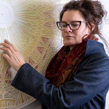 A woman with brown hair and glasses lightly touches a pale coloured artwork with raised sections. She has her eyes closed.