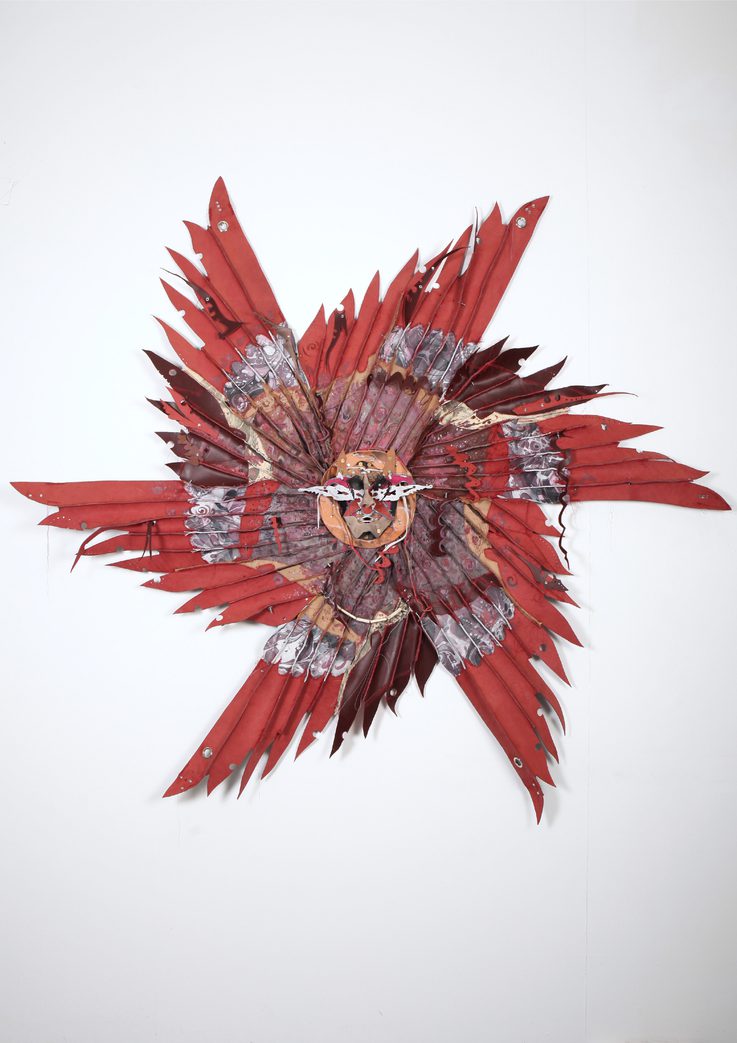A vibrant, abstract wall sculpture featuring a central face surrounded by an array of red and brown feathers. The piece combines intricate patterns and textures, creating a striking visual impact against a plain white background.